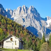 Refuge blanc avec croix en bois niché dans une forêt automnale sous les pics calcaires spectaculaires des Dolomites de Brenta par ciel bleu.