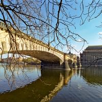 Pont en pierre avec arches enjambant la Vltava à Prague, avec le théâtre national visible sur la rive droite sous des arbres nus d'hiver.