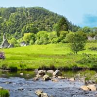 Rivière rocheuse traversant des prairies vertes à Wicklow, Irlande, avec ruines d'église et tour ronde sur la colline, montagne boisée en arrière-plan.