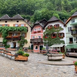 Place pavée à Hallstatt avec maisons alpines traditionnelles aux couleurs jaune, rose et blanche. Bancs en bois, jardinières et fontaine décorent la place.