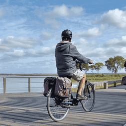 Deux cyclistes avec sacoches roulent sur une passerelle en bois le long d'une baie. Des arbres bordent l'horizon sous un ciel partiellement nuageux en Baie de Somme.