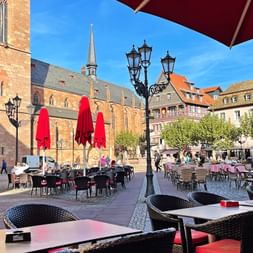 Terrasse de café sur place de marché Palatinat Terrasse de café avec tables et chaises sur une place pavée du Palatinat, avec une église gothique et des bâtiments historiques colorés.