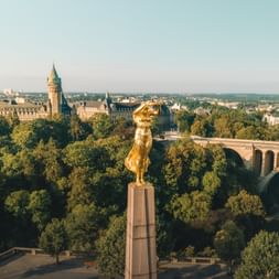 Statue dorée sur colonne de pierre surplombant Luxembourg-Ville avec bâtiments historiques, arbres verts et pont en pierre en arrière-plan.