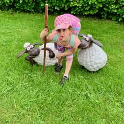 Jeune fille en foulard rose tenant un bâton de berger en bois, posant avec deux moutons décoratifs sur pelouse verte.