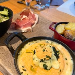 Fromage à raclette fondu dans une poêle en fonte avec herbes, servi avec pommes de terre rôties, tranches de jambon cru et salade verte sur une table en bois.