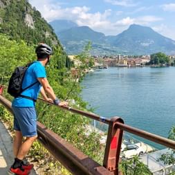 Cycliste en maillot bleu et casque debout à la balustrade surplombant le Lac de Garde avec montagnes et ville italienne en arrière-plan.