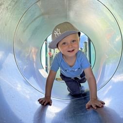 Jeune enfant portant une casquette et un t-shirt bleu rampant dans un tunnel de jeu métallique dans le Salzkammergut lors d'une randonnée familiale.