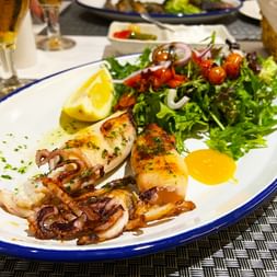 Poulpe grillé servi sur assiette blanche à bord bleu dans restaurant de Majorque, garni de salade de roquette, quartier de citron et huile d'olive.