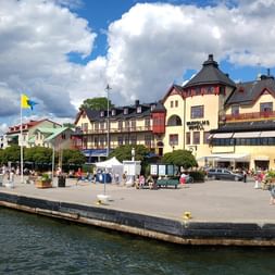 Place au bord de l'eau à Vaxholm avec des gens se détendant par une journée ensoleillée. Bâtiments historiques colorés bordant le port sous un ciel bleu.