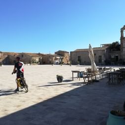 Cycliste avec vélo traversant une grande place pavée à Marzamemi, Sicile. Bâtiments historiques et église entourent la place sous ciel bleu.