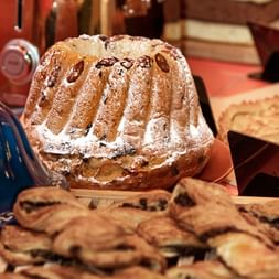 Kougelhof alsacien traditionnel saupoudré de sucre glace sur planche en bois, entouré de viennoiseries et moules colorés.