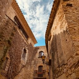 Ruelle médiévale étroite à Pals avec bâtiments en pierre patinée, toits en terre cuite et passages voûtés sous un ciel bleu avec nuages blancs.