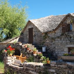 Maison traditionnelle en pierre avec toit d'ardoise aux Gorges du Tarn et Jonte. Pots de fleurs décorent les murs et la terrasse sous ciel bleu.