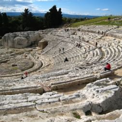 Ruines de théâtre grec antique à Syracuse avec gradins en pierre semi-circulaires taillés dans la colline, visiteurs explorant le site sous ciel bleu.