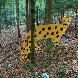 Sculpture de léopard en bois jaune avec des taches noires positionnée entre les arbres dans une forêt. La sculpture se dresse sur un sol forestier couvert de feuilles mortes.