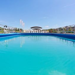 Grande piscine turquoise sur le pont du navire de croisière MS Manon avec garde-corps métalliques, ciel bleu et bâtiments distants.