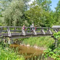 Deux cyclistes avec vélos traversent un pont en bois au-dessus d'un petit ruisseau. Végétation verte dense et arbres entourent le cours d'eau.