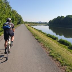 Cycliste roulant sur chemin asphalté le long d'un canal calme en République tchèque. Arbres verts bordent le côté gauche, eau reflète le ciel.