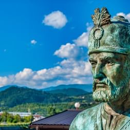 Statue en bronze du Roi Béla IV avec couronne ornée et barbe contre ciel bleu avec montagnes et bâtiments en arrière-plan à Visegrad.