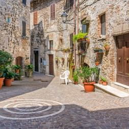 Ruelle pavée étroite à Montecchio avec motif circulaire en pierre, bâtiments anciens, volets en bois et plantes en pot décorant les murs.