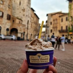 Main tenant une coupe de glace Gelateria Dondoli sur la place médiévale de San Gimignano avec bâtiments en pierre et touristes en arrière-plan.