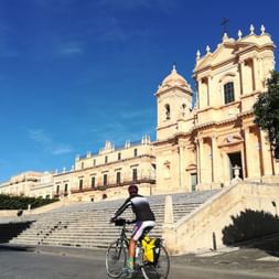 Cycliste avec sacoches jaunes devant la cathédrale baroque de Noto en Sicile. L'église en calcaire doré présente deux clochers et une façade ornée.