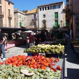 Marché en plein air en Costa Brava avec des produits frais. Poivrons rouges, artichauts et melons au premier plan. Bâtiments historiques.