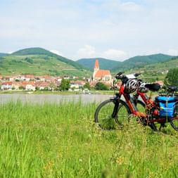Deux vélos de randonnée avec sacoches garés dans les hautes herbes près du Danube, avec ville autrichienne et clocher en arrière-plan.