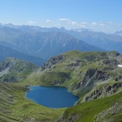 Lac bleu de montagne entouré de prairies alpines vertes et de pics rocheux dans le Queyras, avec des montagnes enneigées au loin.