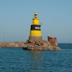 Phare jaune et noir marqué 'MEN' sur île rocheuse dans les Côtes-d'Armor, Bretagne. Mer bleue entoure rochers brun-rouge, voiliers visibles au loin.