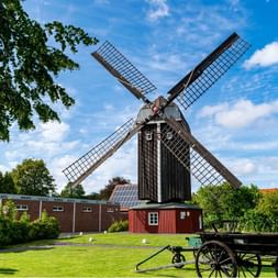 Moulin à vent traditionnel en bois à Dornum avec quatre ailes à treillis contre le ciel bleu. Pelouse verte avec vieille charrette, bâtiment rouge.