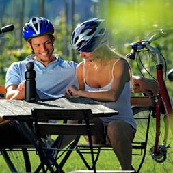 Cyclistes se reposant sur piste Neckar Deux cyclistes casqués assis à table en bois lors d'une pause sur piste cyclable du Neckar, vélos garés dans cadre forestier ensoleillé.