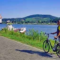 Cycliste avec vélo sur chemin pavé au bord du Rhin. Bateau à passagers blanc amarré, collines et ville en arrière-plan.