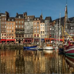 Bâtiments historiques colorés bordent le port d'Honfleur avec des bateaux à voile traditionnels amarrés dans l'eau calme reflétant l'architecture.