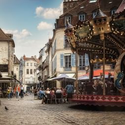 Manège traditionnel sur une place pavée à Dijon, Bourgogne, avec des bâtiments historiques et des cafés en terrasse sous un ciel bleu.