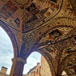 Plafond voûté orné de fresques Renaissance à Sienne, avec des scènes religieuses et motifs décoratifs. Bâtiments médiévaux en brique visibles à travers les arches.