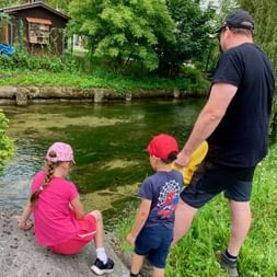Père avec deux enfants pêchant près d'une rivière claire à Bad Goisern. Fille en rose assise sur la berge, garçon en casquette rouge debout.