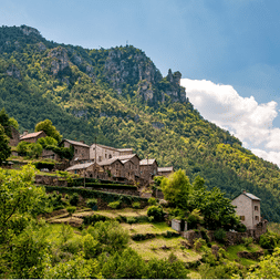 Village traditionnel en pierre sur une colline des Cévennes avec des pentes en terrasses, entouré de forêts vertes et de sommets rocheux sous un ciel bleu.