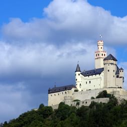 Le château de Marksburg perché sur une colline boisée sous un ciel bleu avec des nuages blancs. La forteresse médiévale a des murs blancs.