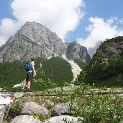 Randonneur avec sac à dos bleu sur pont en bois face aux pics rocheux des Dolomites de Brenta avec pentes vertes et ciel bleu nuageux.