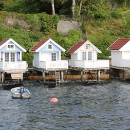 Quatre cabanes de bateaux en bois blanc avec toits rouges sur pilotis au-dessus de l'eau du fjord d'Oslo. Un petit bateau à moteur flotte à proximité.