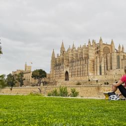 Cycliste en maillot rose assise sur l'herbe avec casque de vélo à côté, face à la cathédrale gothique de Palma à Majorque avec palmiers.
