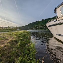 Bateau de croisière blanc MS Olympia amarré sur la Moselle avec des collines vertes et vignobles en arrière-plan sous un ciel nuageux.