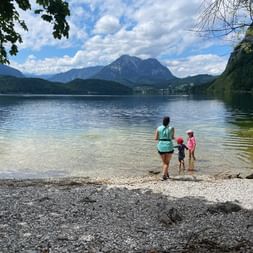 Femme avec deux enfants dans l'eau peu profonde au bord du lac Altaussee. Montagnes et ciel nuageux en arrière-plan, plage de galets au premier plan.