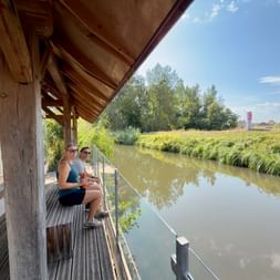 Deux personnes assises sur une terrasse en bois surplombant un canal calme. Végétation verte le long des rives, maisons visibles au loin sous ciel bleu.