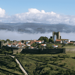 Vue aérienne de la ville fortifiée médiévale de Bragança avec tour de château et maisons aux toits rouges, entourée de champs verts et montagnes brumeuses.