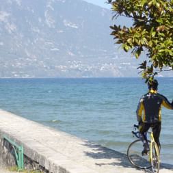 Cycliste avec vélo debout sur le sentier en béton au bord du lac d'Annecy, avec montagnes et eau bleue en arrière-plan.