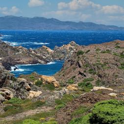 Côte rocheuse escarpée au Cap de Creus avec végétation verte, mer Méditerranée bleue profonde et montagnes lointaines sous un ciel nuageux.