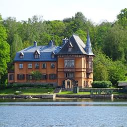 Bâtiment orange avec toit bleu et tour au bord de l'eau à Skansen, entouré d'arbres verts. Jetée en pierre dans l'eau calme.
