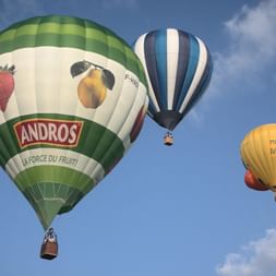 Trois montgolfières colorées flottant dans le ciel bleu au-dessus de la Dordogne. Le ballon le plus proche affiche la marque Andros avec fraise et pêche.